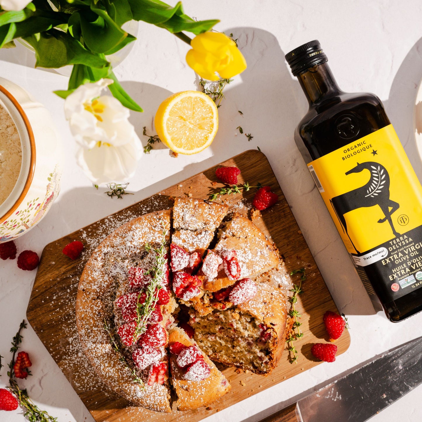Top-down photo of a freshly baked raspberry thyme olive oil cake sliced on a wooden board, surrounded by raspberries, lemon, and a bottle of Terra Delyssa Organic Extra Virgin Olive Oil.