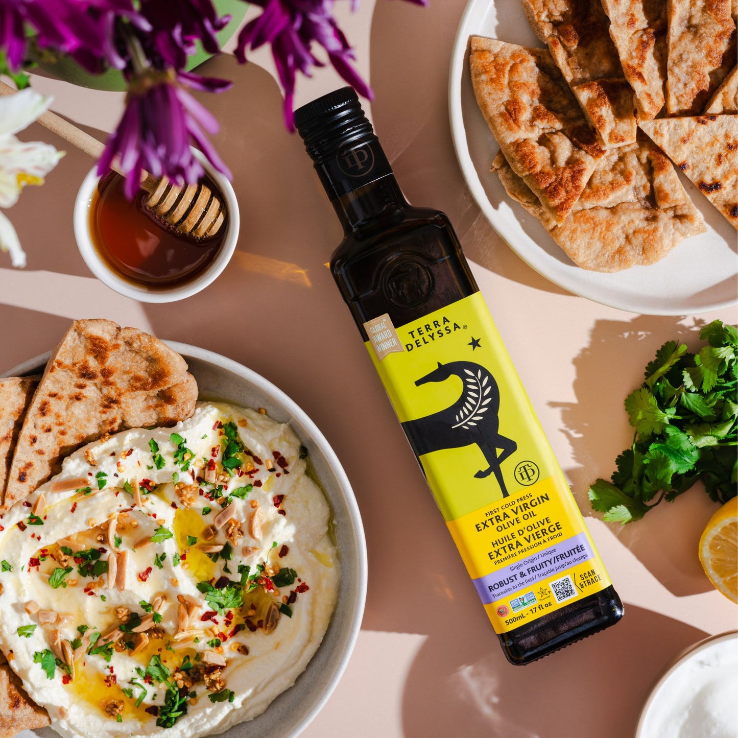 Top-down view of Terra Delyssa Extra Virgin Olive Oil with flatbread, whipped feta topped with herbs, nuts, and spices, accompanied by honey and fresh cilantro on a soft pink background with flowers in the corner.
