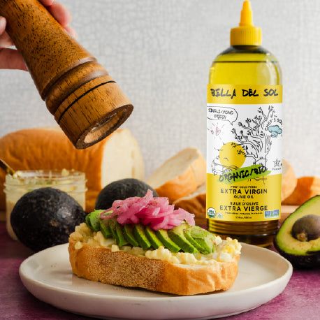 
                  
                    Bella Del Sol Organic Extra Virgin Olive Oil in a chef squeeze bottle beside avocado toast with pickled onions, fresh bread, and pepper grinder – healthy breakfast scene
                  
                