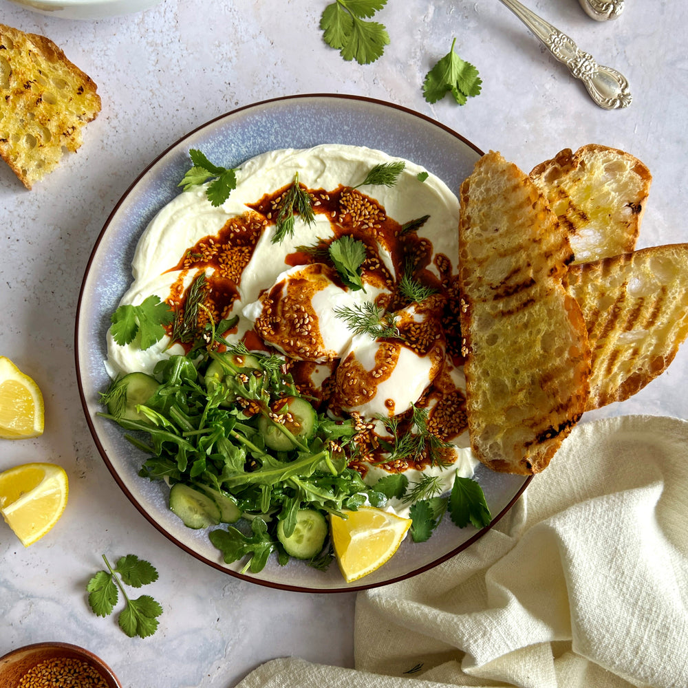 A scrumptious image of a traditional Turkish egg dish featuring perfectly poached eggs nestled atop a bed of creamy and garlicy yogurt, enhanced with a luscious Terra Delyssa Extra Virgin Olive Oil-infused chili, and sprinkled with fresh herbs